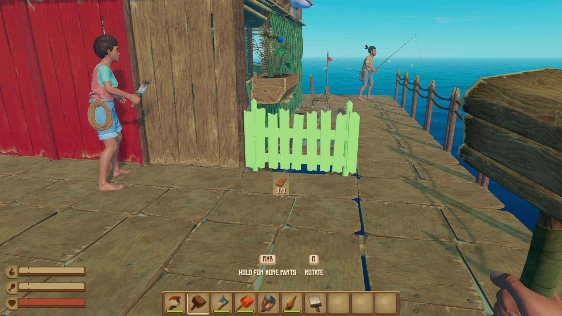 Raft-Spielszene mit grüner Zaun-Bauvorschau neben einer Holzhütte, während andere Spieler streichen und angeln; Bauhammer und Überlebensanzeigen sind sichtbar.