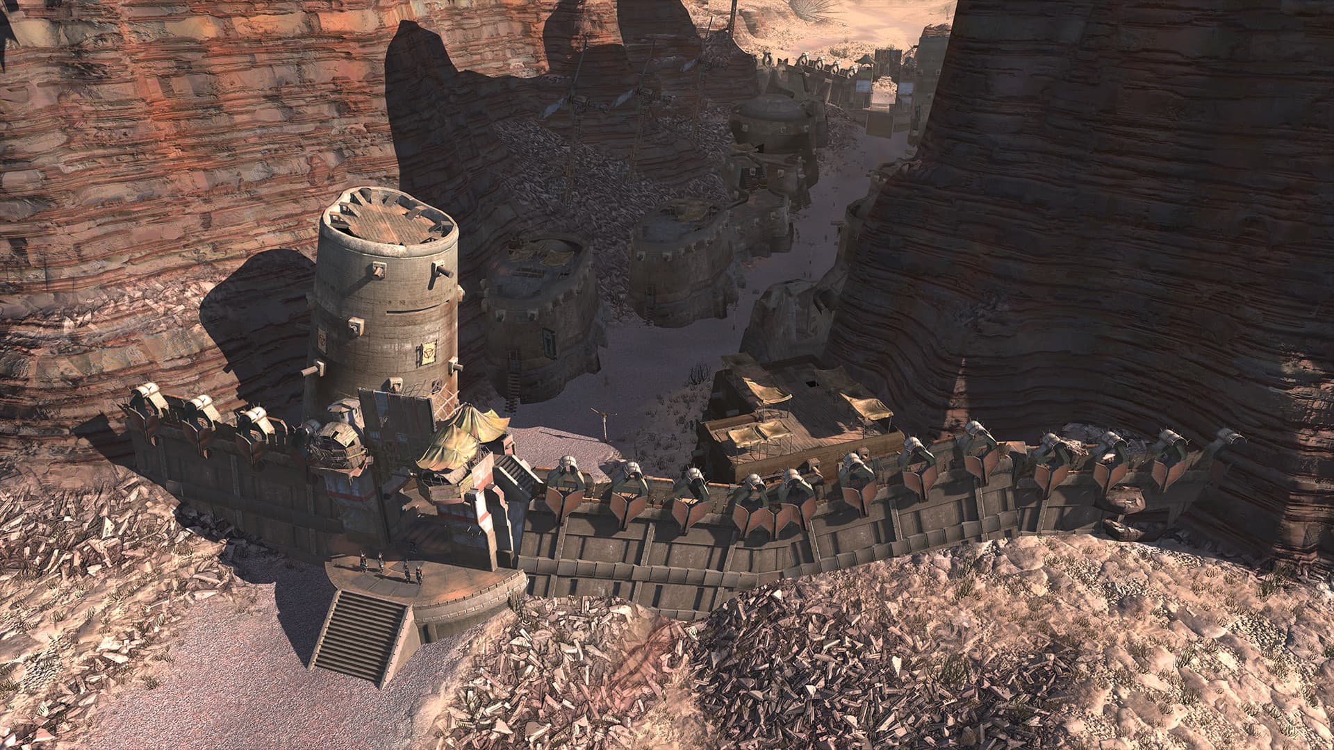 Kenshi in-game view of a fortified canyon settlement with towers, buildings and figures near the entrance.