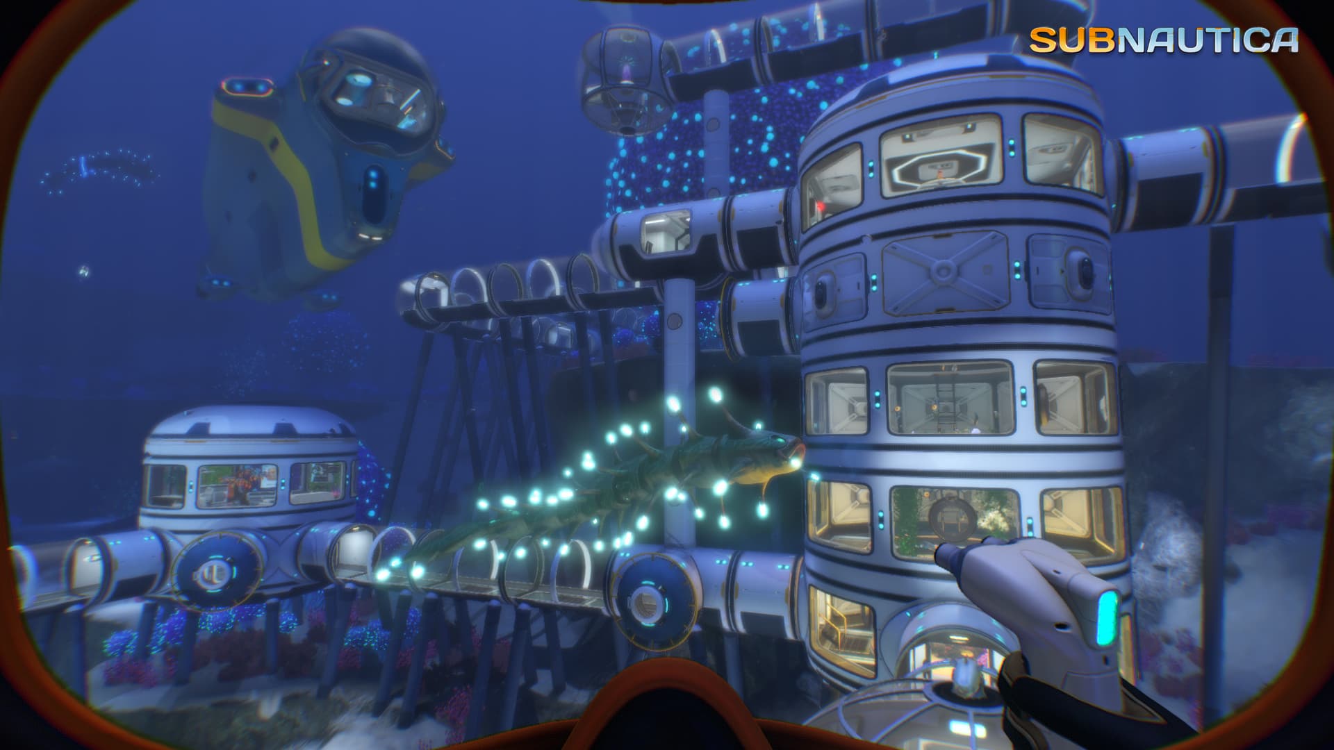 Subnautica aus der Ego-Perspektive: ein beleuchtetes mehrstöckiges Unterwasserhabitat mit Fenstern, Verbindungsgängen und einem Tauchfahrzeug in der Nähe.